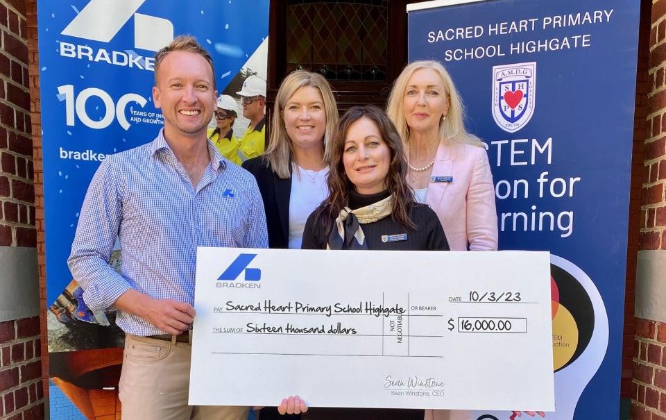 Four people, one man and three women stand holding a novelty cheque in front of banners displaying the Bradken and Sacred Heart Primary School logos.